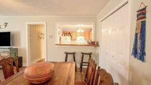 Un área de comedor con una mesa de madera, sillas y un otomano decorativo. Se ve una cocina a través de un arco abierto.