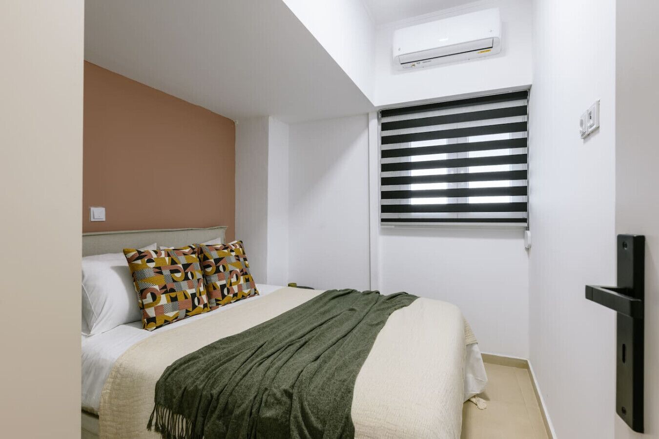 A bedroom with a bed, a patterned pillow, a green throw blanket, a window with blinds, and an air conditioning unit.
