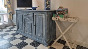 Una habitació amb un sòl de quadradets, un armari gris amb calaixos, una televisió de pantalla plana, un ram de flors decoratiu i una taula petita blanca amb llibres.