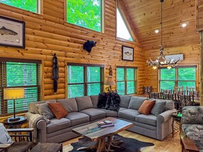 A log cabin interior with a large L-shaped sectional sofa, a chandelier, and a dining area with chairs.