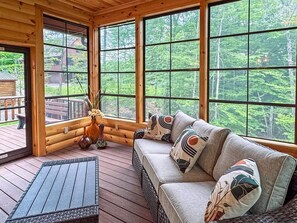 A wooden deck with a sofa and a table, large windows with a forest view, and a deer head decoration.