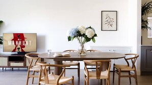 A dining area with a round table, six wicker chairs, a vase with flowers, a framed picture on the wall, and a TV displaying 'MANHUNT' on the left. - Luxurious 3-Bedroom Notting Hill (Greater London)