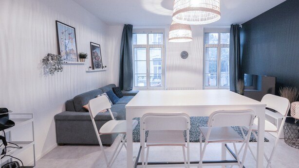 A modern living room with a large white table, grey sofa, and white chairs. There are two windows with curtains, a chandelier, and wall-mounted artwork. - Urban Calm - Bright & Modern Stay (Le Mans)