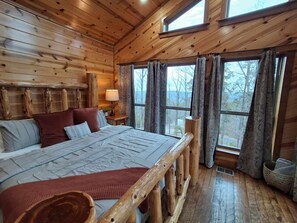 Un dormitorio de cabaña de madera con una cama grande, una mesita de noche y una ventana con cortinas.