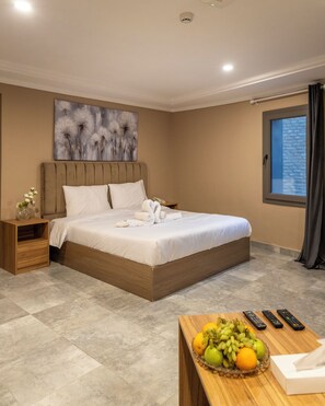 Une chambre d’hôtel avec un lit, une table de chevet ornée d’un vase, un panier de fruits et une fenêtre avec des rideaux.