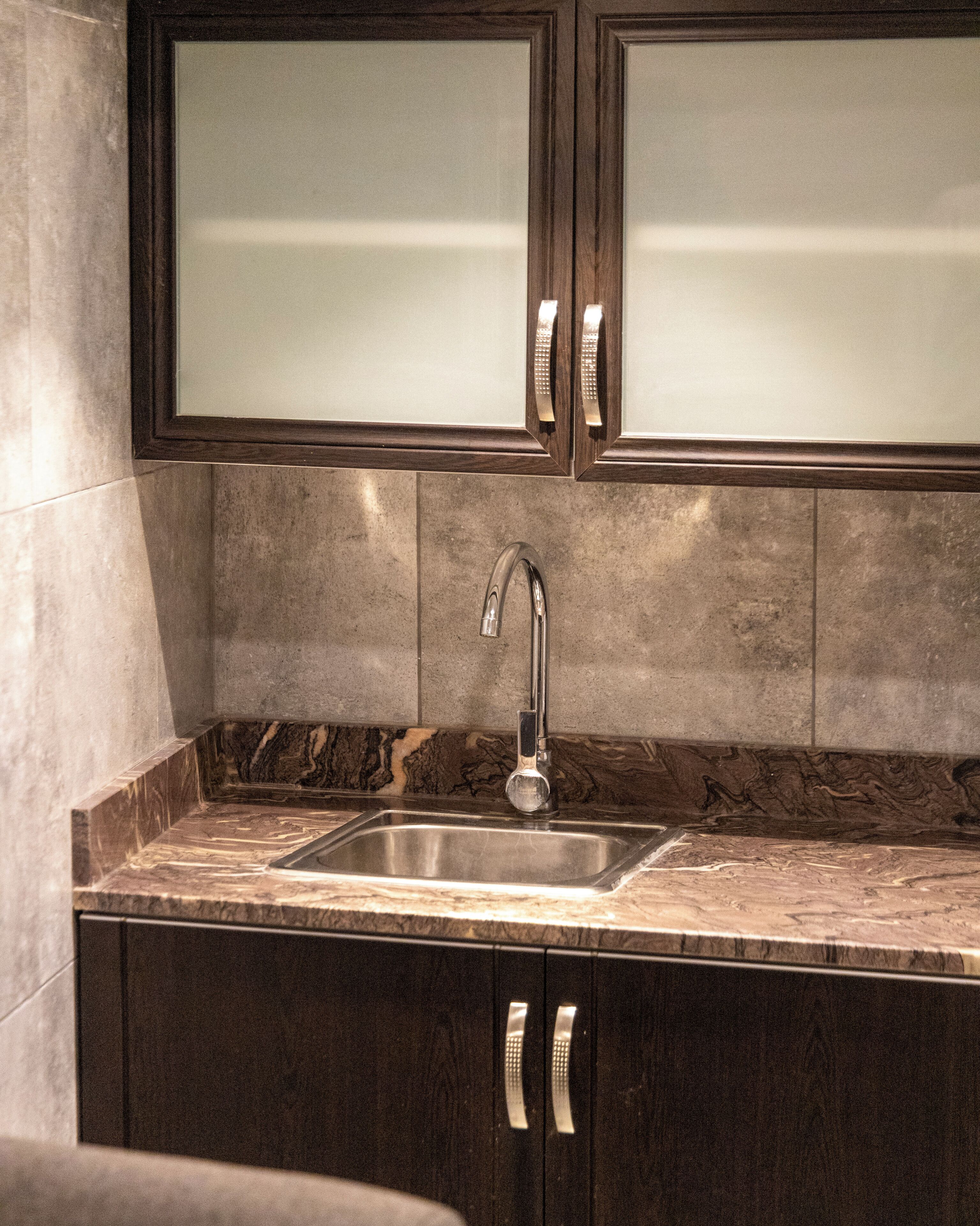 A kitchen with a stainless steel sink, dark wooden cabinets with frosted glass doors, and a marble countertop.