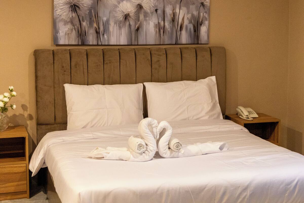 Hotellihuone, jossa on suuri sänky, puinen pääty, valkoiset vuodevaatteet, sängyn yläpuolella oleva taulu, yöpöytä, jossa on puhelin, ja maljakko, jossa on kukkia.