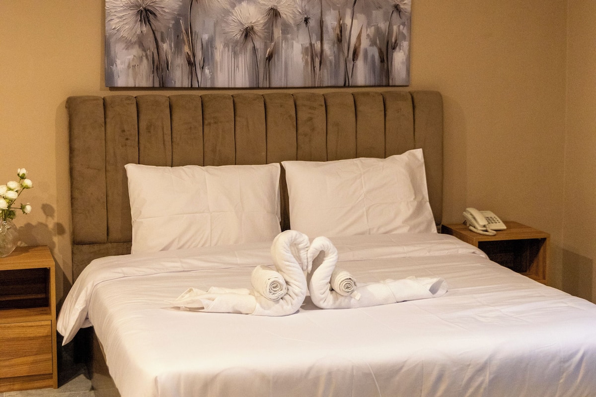 Habitación de hotel con una cama grande, cabecera de madera, ropa de cama blanca, un cuadro sobre la cama, una mesita de noche con un teléfono y un jarrón con flores.
