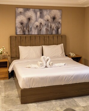 Une chambre d’hôtel avec un grand lit, une tête de lit en bois, du linge de lit blanc, un tableau au-dessus du lit, une table de chevet avec un téléphone et un vase rempli de fleurs.