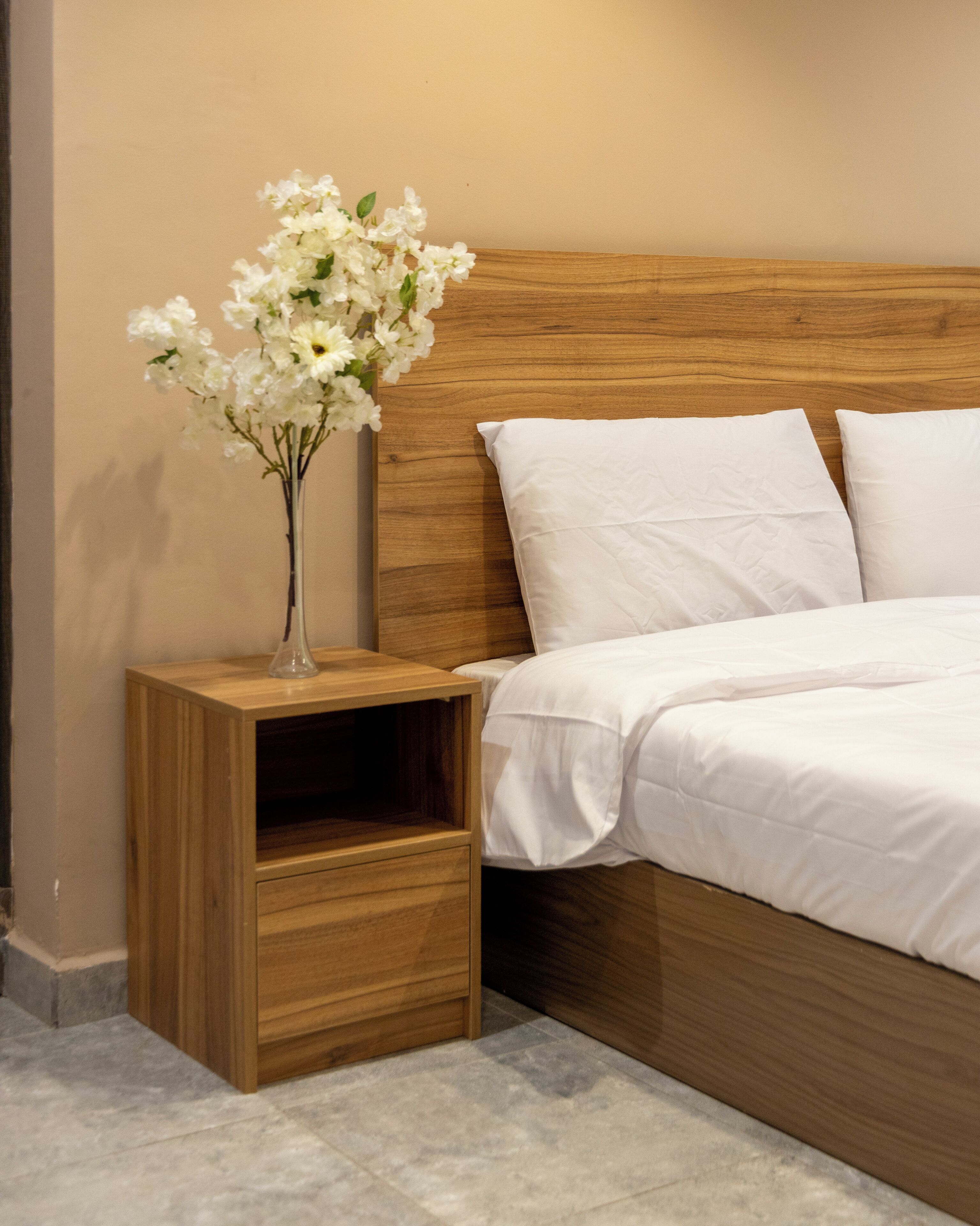 A bedroom with a wooden headboard, white bedding, a nightstand with a vase of flowers, and a small wooden cabinet.