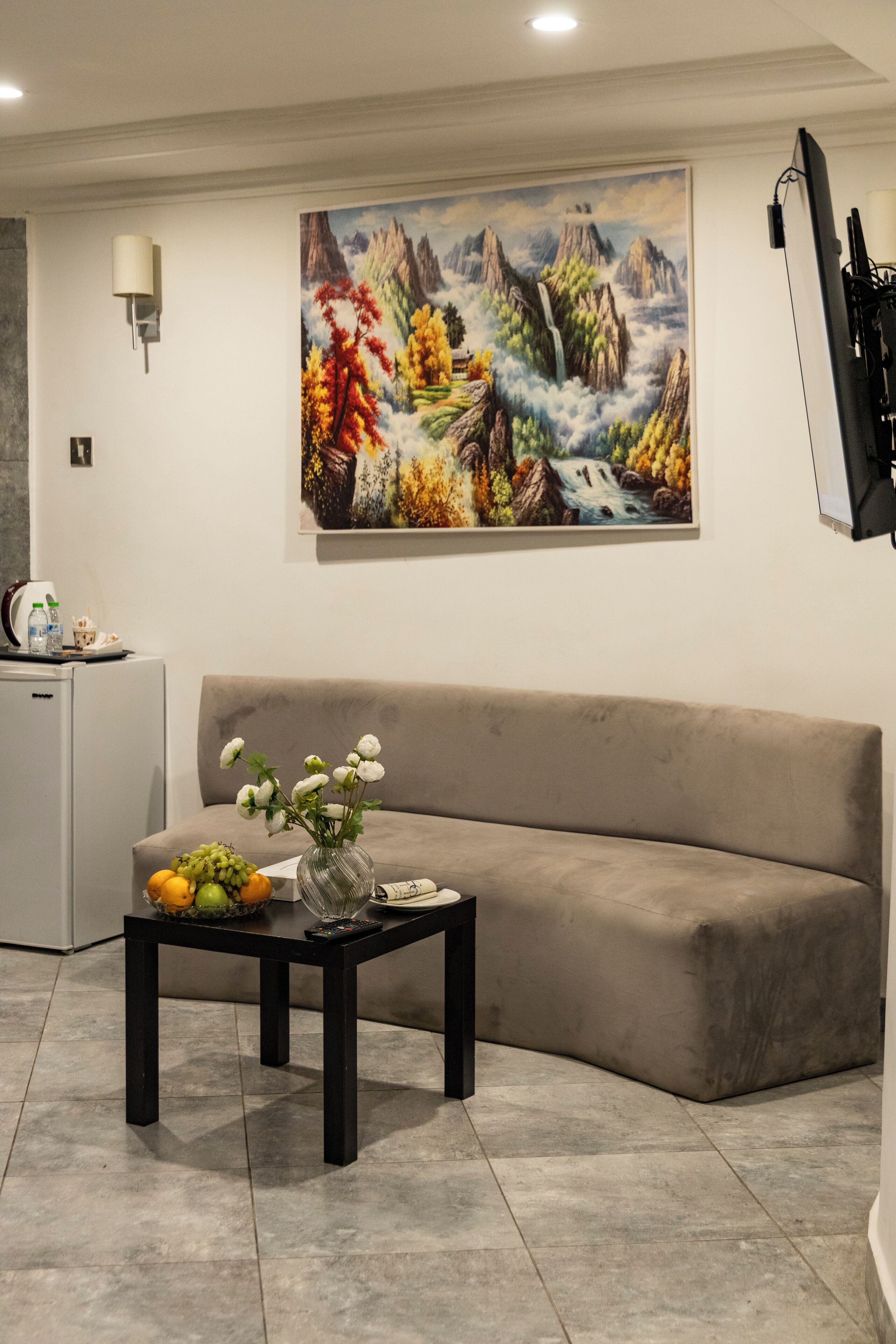 A modern living room with a beige sofa, a black coffee table with a vase of flowers, a painting of a mountainous landscape, and a flat-screen TV mounted on the wall.