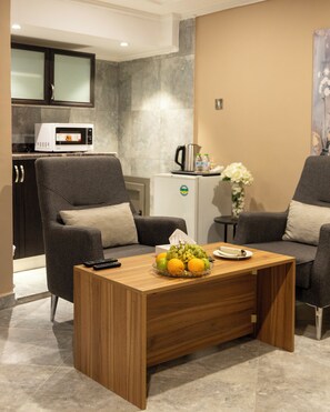 Un salon moderne avec une table basse en bois, deux fauteuils gris, un petit coin cuisine équipé d’un four à micro-ondes et d’un réfrigérateur.