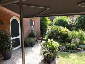 Un patio cubierto con pared de ladrillo, plantas en macetas, una silla de madera y un jardín con arbustos y flores variadas.