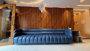 A modern living room with a blue leather sofa, wooden wall paneling, and a glass door leading to another room. - Hotel Anu  (Phulpur)