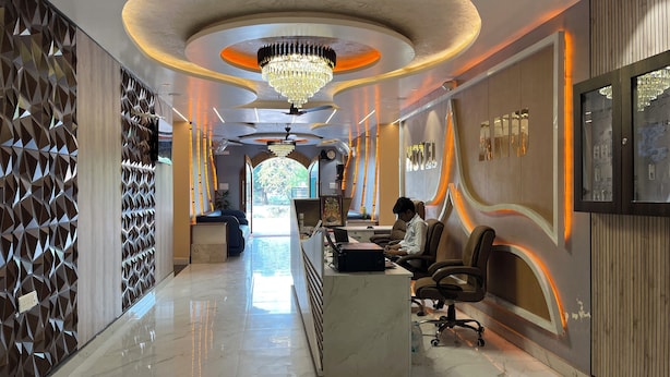 A hotel reception area with a modern design, featuring a chandelier, a reception desk, and a seating area with a view of the outdoors. - Hotel Anu  (Phulpur)
