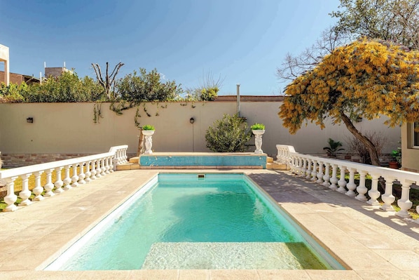 A rectangular swimming pool with a white balustrade, surrounded by a beige wall and greenery.