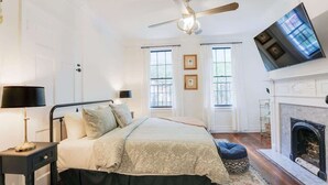 A bedroom with a bed, a ceiling fan, a fireplace, and a TV mounted on the wall. - Charming Historic Apt Steps to River St & Tours (Savannah)