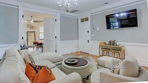 A living room with a sofa, ottoman, and armchairs, a chandelier, a TV mounted on the wall, and a dining area with a table and chairs.