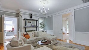 A living room with a sectional sofa, ottoman, chandelier, fireplace, and a view into another room.