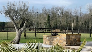 Una jardinera de piedra con un pequeño arbusto, rodeada por una terraza de madera y una zona ajardinada con un árbol y una valla al fondo.
