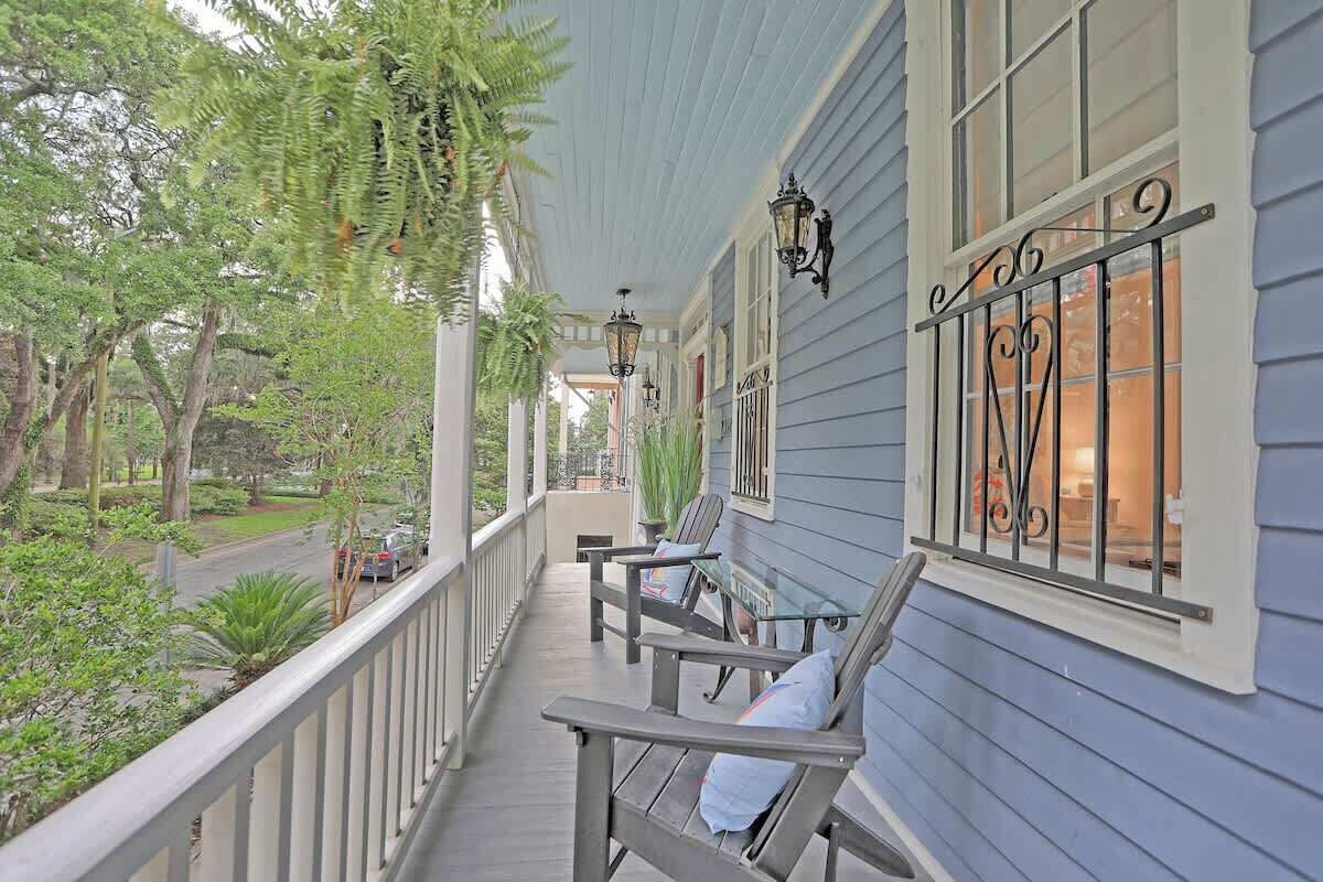 A covered porch with wooden chairs, a railing, and a view of a street and greenery.