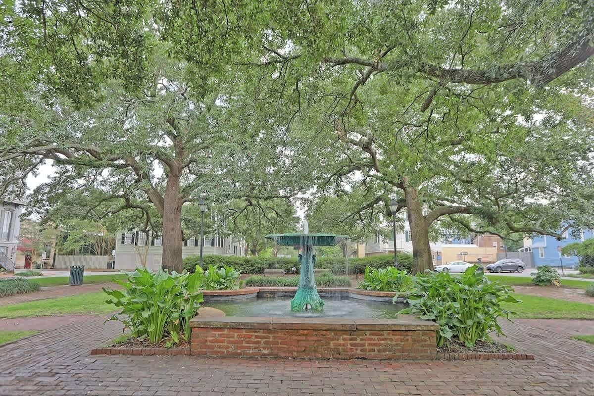 A fountain surrounded by brickwork, with trees and a paved walkway.