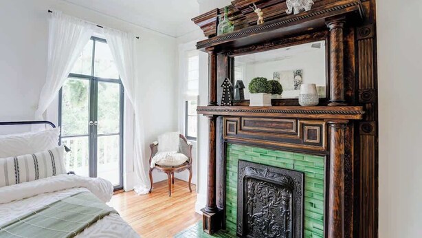 A room with a bed, a chair, a fireplace with a green brick surround, and a large window with white curtains.