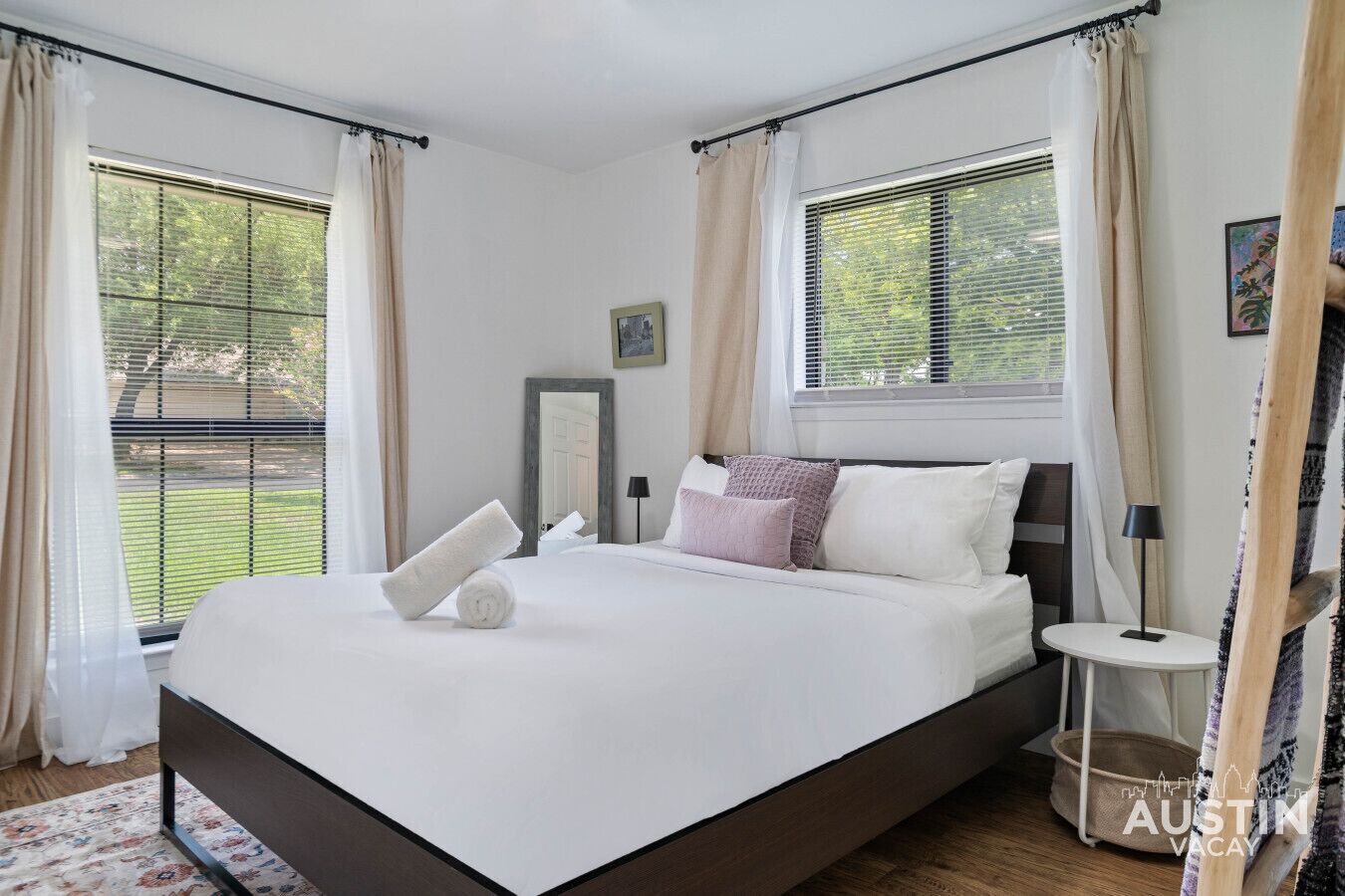 A bedroom with a large bed, two windows with blinds, a bedside table with a lamp, and a framed picture on the wall.