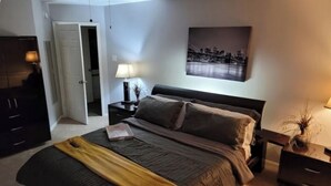 Un dormitorio con cama, mesita de noche, lámpara, un cuadro y una puerta que da a otra habitación.