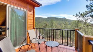 Terraza de madera con dos sillas y una mesita, con vistas a una montaña boscosa.