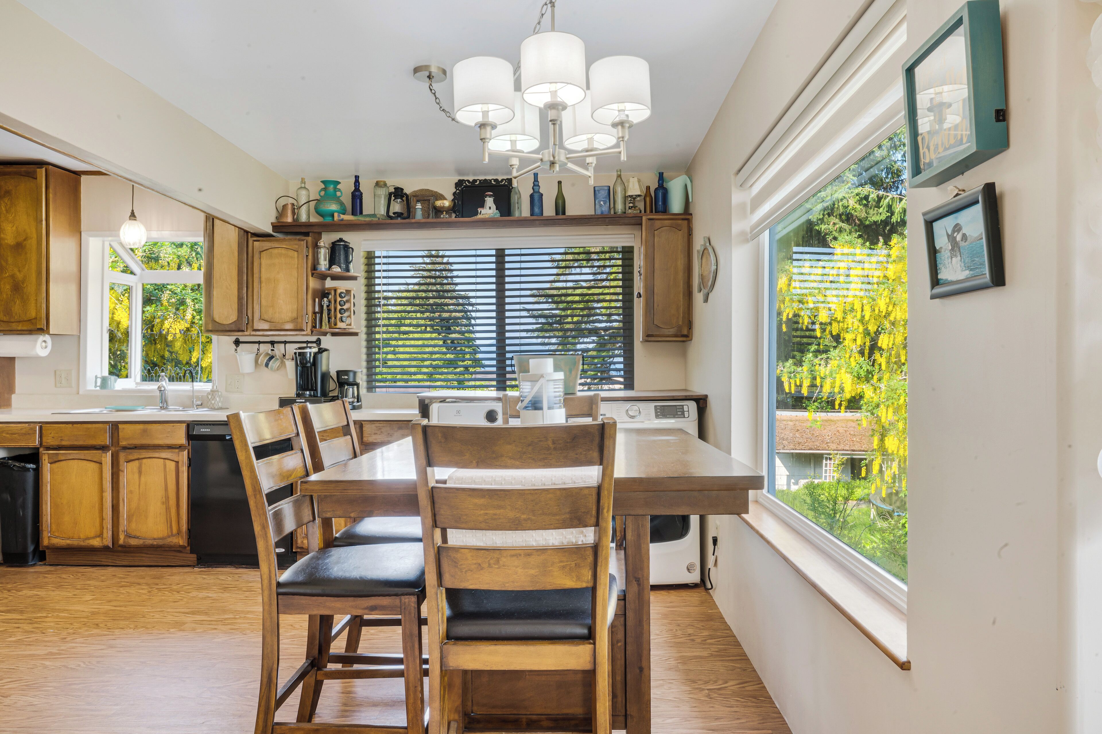 Une cuisine avec des meubles en bois, une table à manger avec des chaises, un lustre et une grande fenêtre donnant sur un paysage verdoyant.