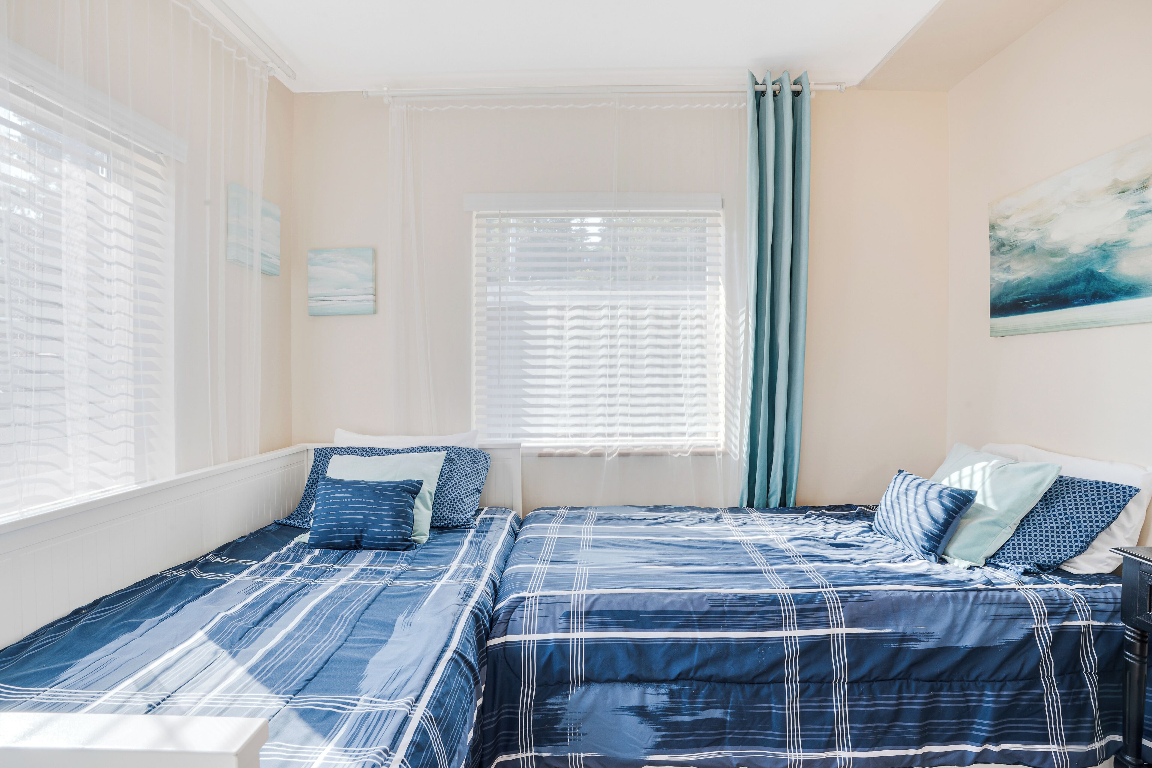 Une chambre avec deux lits, chacun agrémenté de draps bleus et d’éléments décoratifs blancs, une fenêtre avec des stores et un tableau au mur.