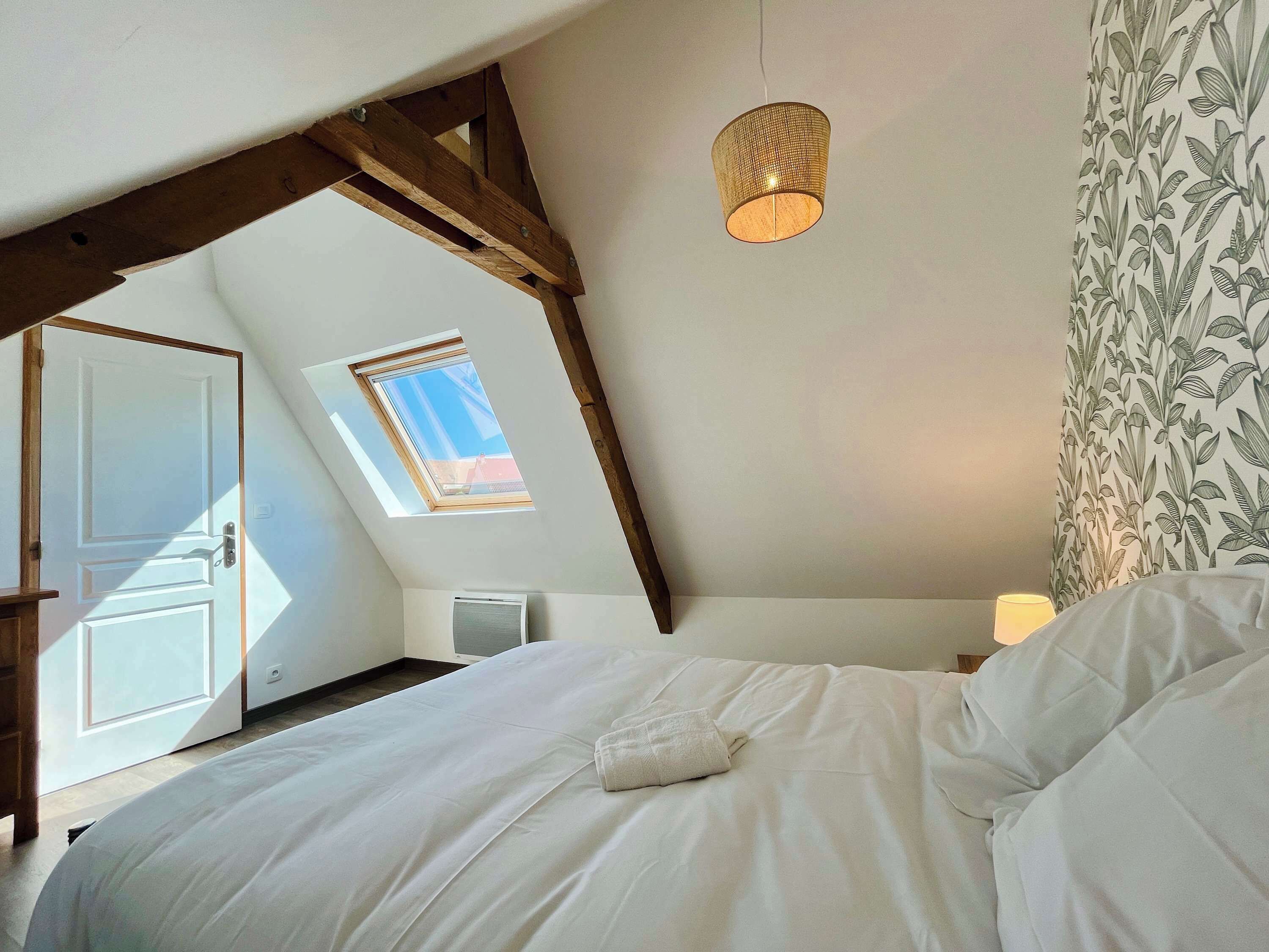 A bedroom with a sloped ceiling, a skylight, a bed with white bedding, a hanging light fixture, and a floral wallpaper pattern.