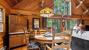 Interior de una cabaña de madera con comedor, armarios de madera y un amplio ventanal con vistas al bosque.