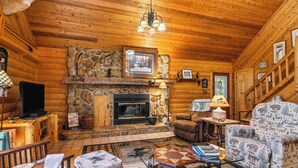 Interior acogedor de una cabaña de troncos con chimenea de piedra, paredes de madera, un televisor y un sillón con estampado.