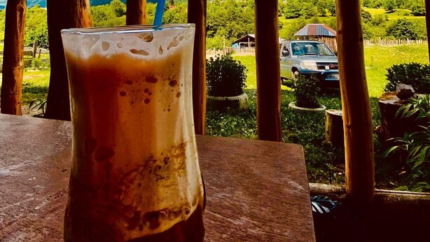 A glass of iced coffee on a wooden table with a view of a mountain landscape and a car in the background. - Qvis sakhli (Mestia)