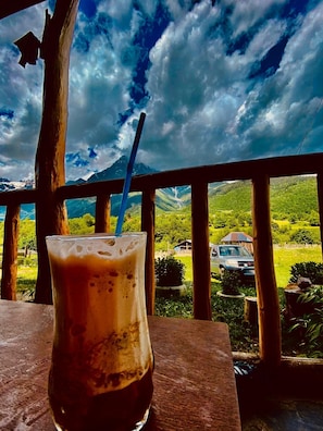 Un verre de café glacé sur une table en bois, avec vue sur un paysage montagneux et une voiture au loin.