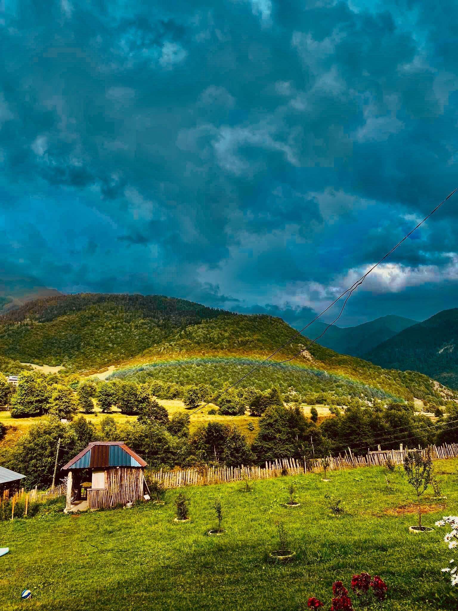 Un paisaje rural con una casa de madera, una zona cercada y un fondo montañoso.