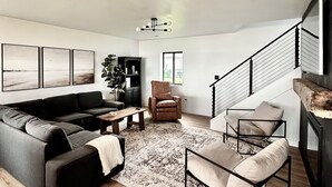 A spacious living room with a black sofa, a wooden coffee table, and a patterned rug. There is a black armchair and a metal-framed chair with cushions. A staircase with a metal railing leads to an upper level.