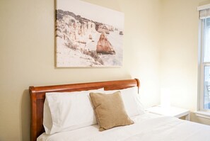 A bedroom with a wooden headboard, white bedding, a beige pillow, and a coastal landscape painting on the wall. - 2 Connected Apartments · 6BR 4BA · Sleeps 12 · W/D (Moss Point)