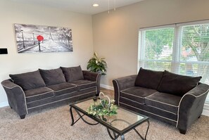 A living room with two dark brown sofas, a glass coffee table with a heart-shaped decoration, and a painting of a street scene on the wall. - 2 Connected Apartments · 6BR 4BA · Sleeps 12 · W/D (Moss Point)