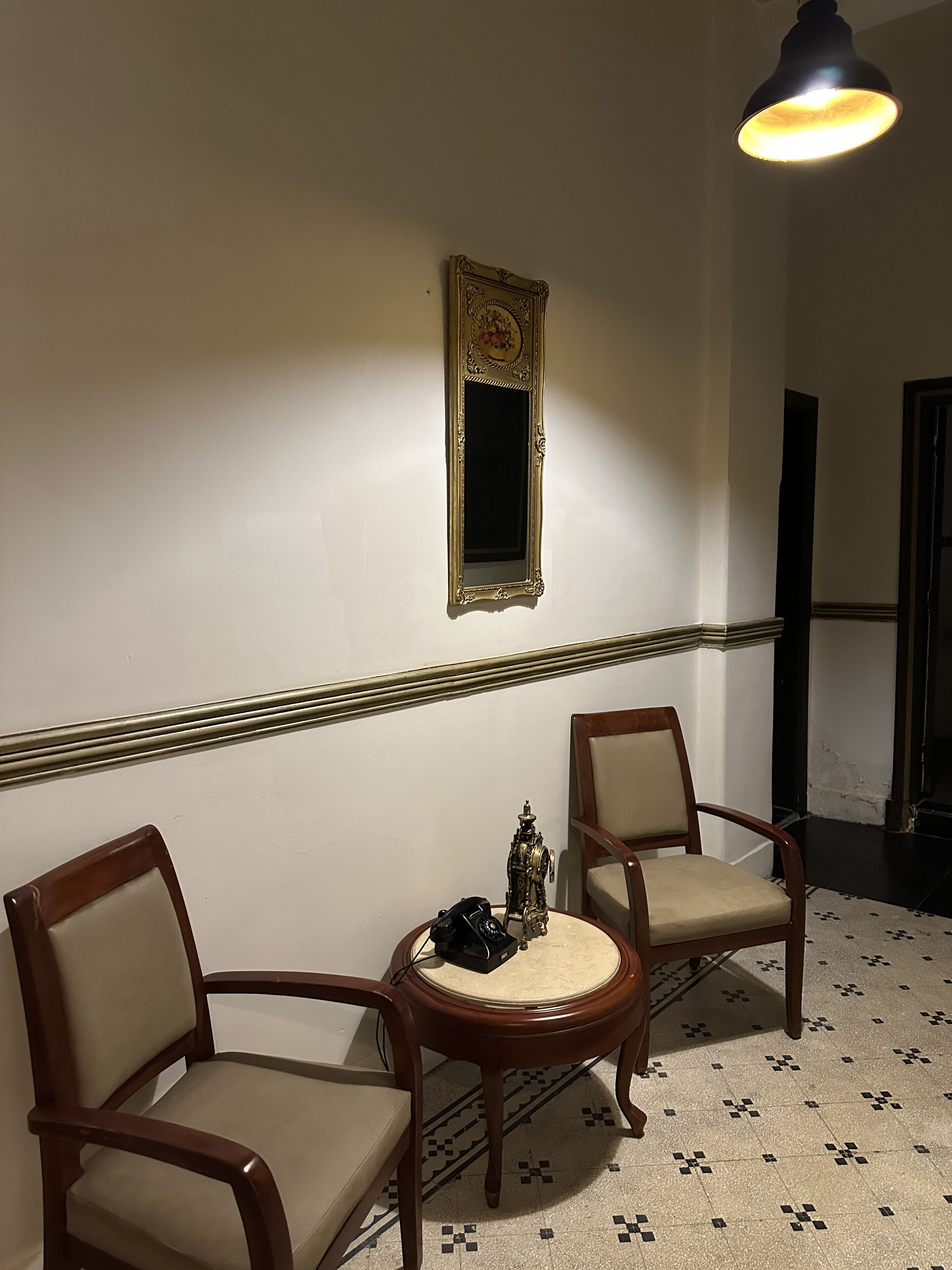 Una habitación con dos sillas de madera, una mesita redonda pequeña, un teléfono antiguo y un espejo decorativo en la pared.