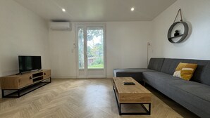 A modern living room with a sofa, a coffee table, a TV on a wooden stand, and a sliding glass door leading to a garden. - Peaceful Home With Terrace & Parking (Le Mans)