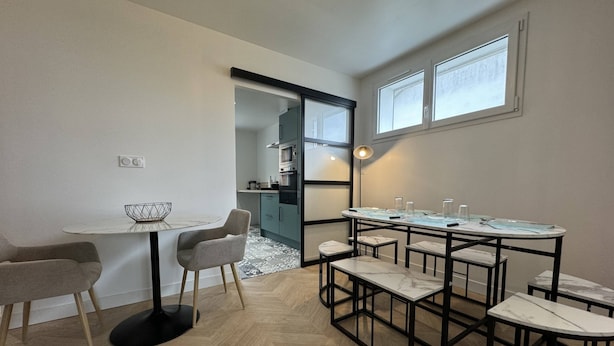 A modern dining area with a herringbone wood floor, a round table with chairs, and a kitchen in the background. - Peaceful Home With Terrace & Parking (Le Mans)