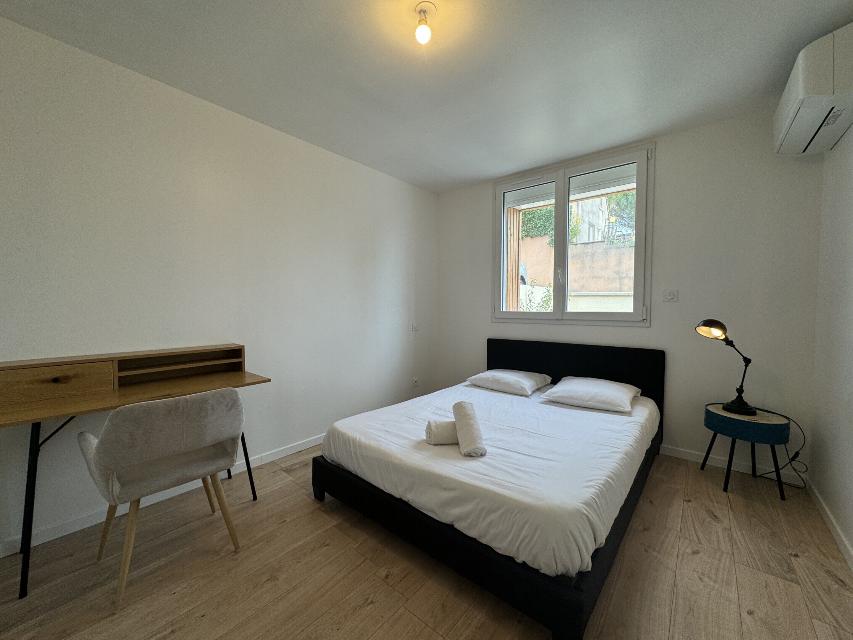 A bedroom with a bed, a desk, and a chair. There is a window with a view of greenery outside.