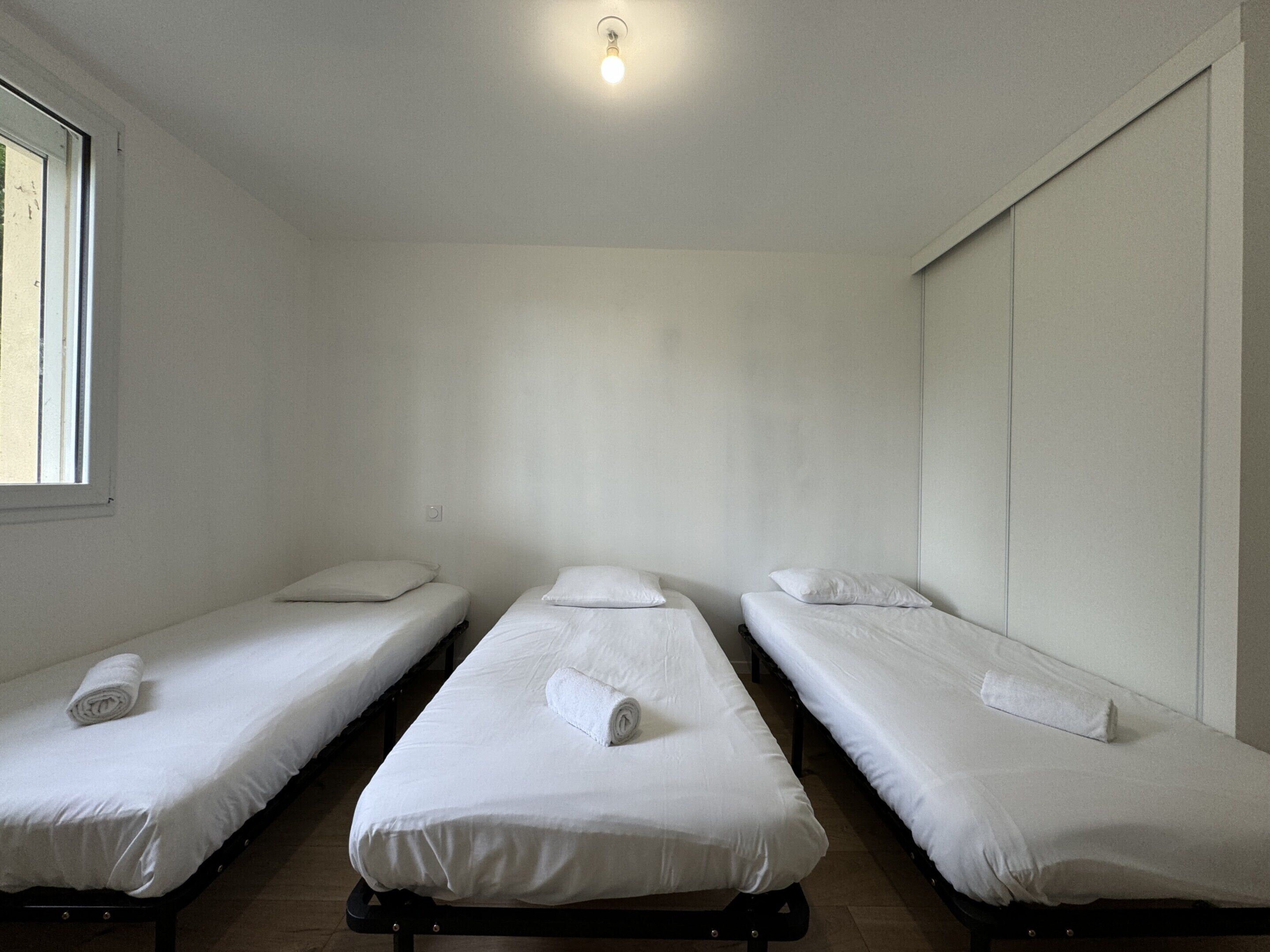 Habitación con tres camas individuales, cada una con una toalla blanca enrollada, una ventana a la izquierda y una lámpara en el techo.