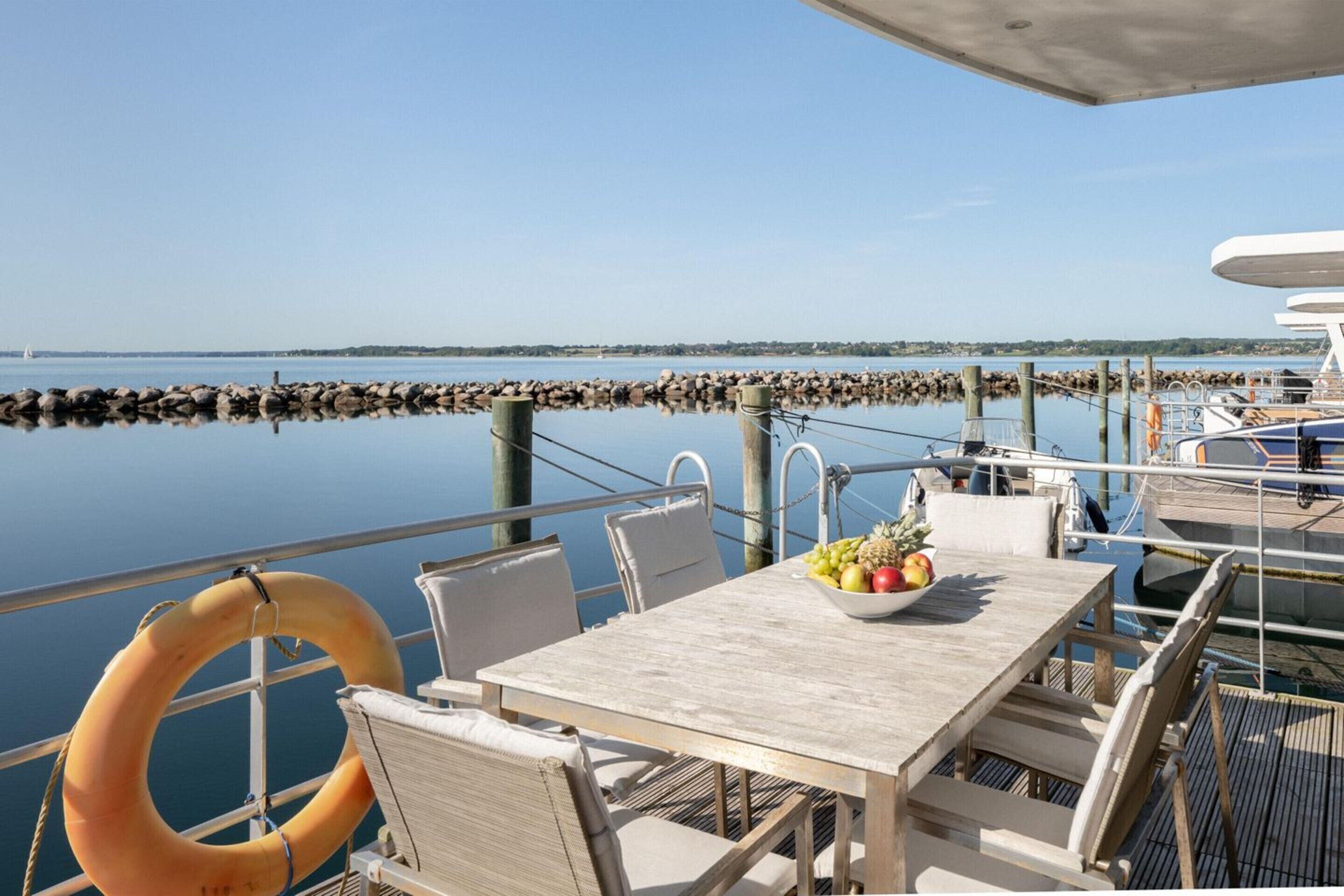 A dining area with a table, chairs, and a bowl of fruit on a dock with a view of the water.