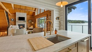 Eine moderne Küche mit Kamin aus Stein, Holzbalken und einem großen Fenster mit Blick auf ein Gewässer.