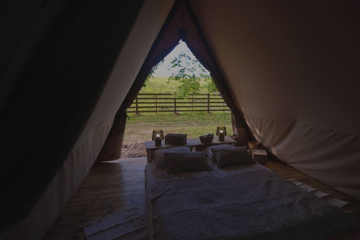 Una tienda de campaña con una cama en su interior, vista a un área verde afuera y una cerca en la distancia.