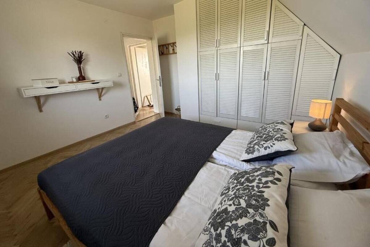 Un dormitorio con cama, un armario, una mesita de noche, una lámpara y una pequeña estantería con un jarrón.